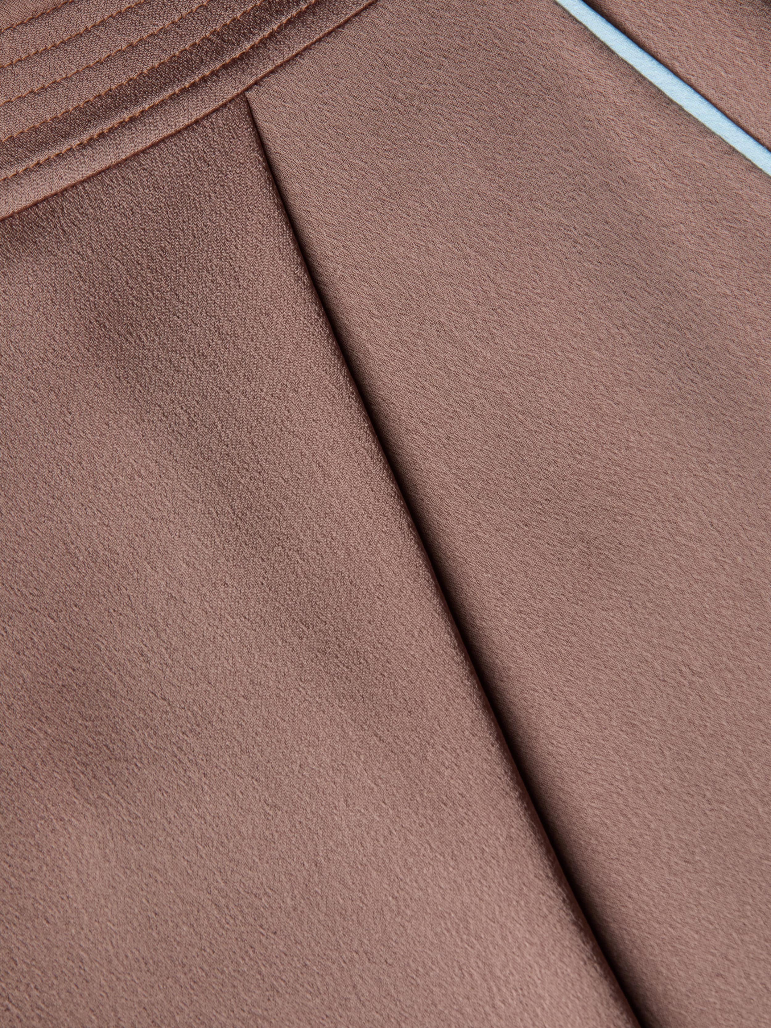 Close-up of a brown textured satin fabric with pleats and a diagonal seam, featuring light blue trim along the edge, as seen on the chic Tavia Satin Trouser.