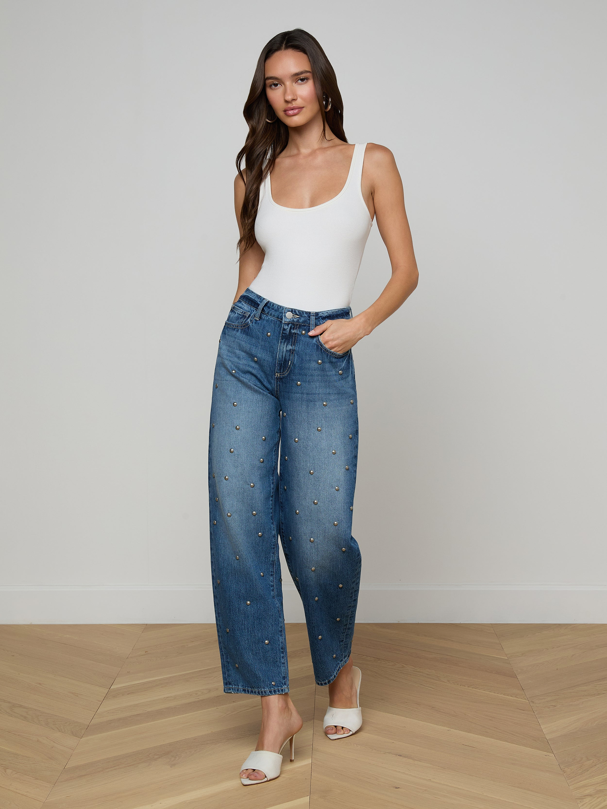 A woman stands against a plain white background wearing a white tank top, the Bex Studded Rodeo Jean in blue high-rise with small studded denim embellishments, and white open-toe heels.