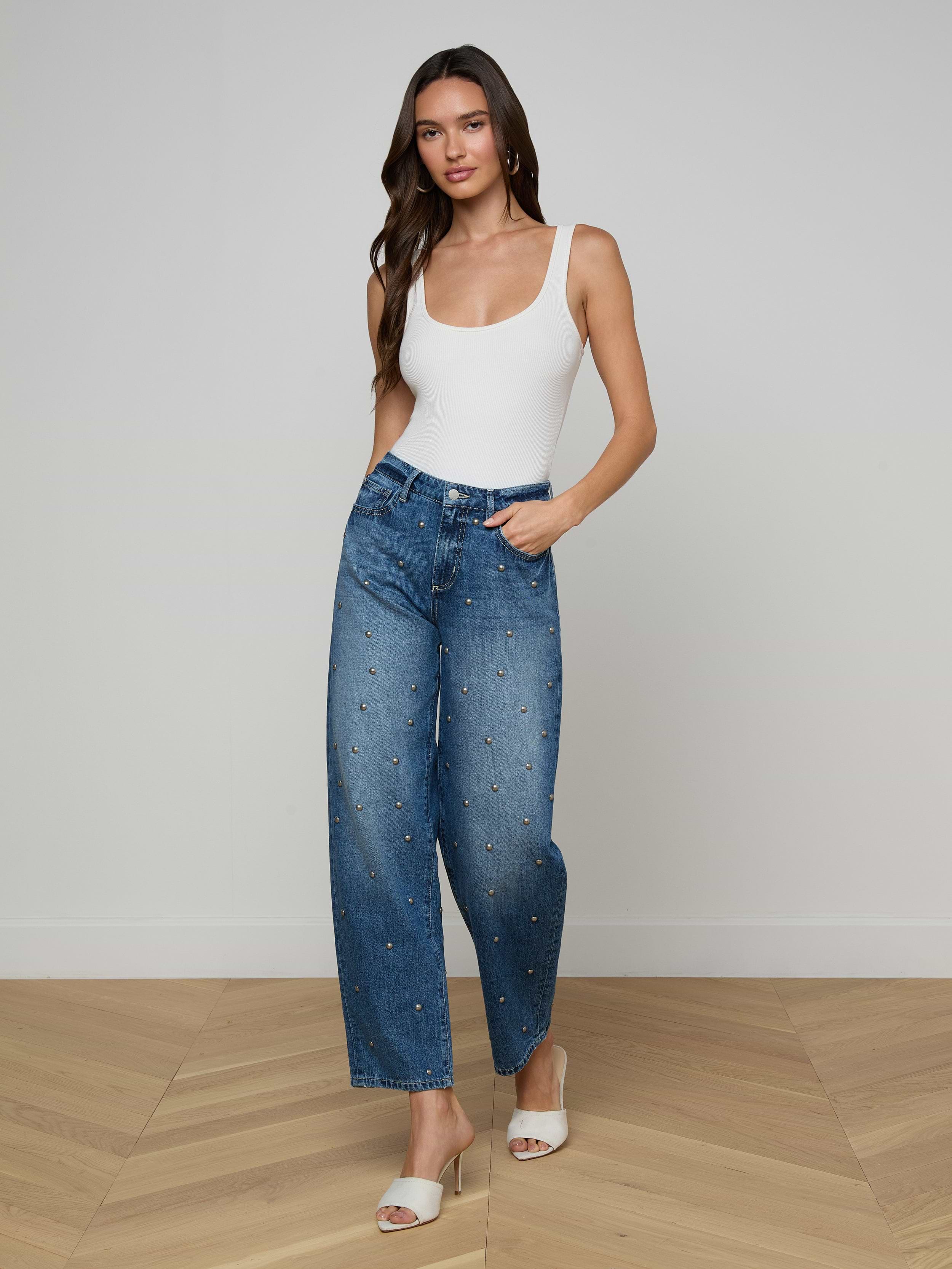 A woman stands against a plain white background wearing a white tank top, the Bex Studded Rodeo Jean in blue high-rise with small studded denim embellishments, and white open-toe heels.