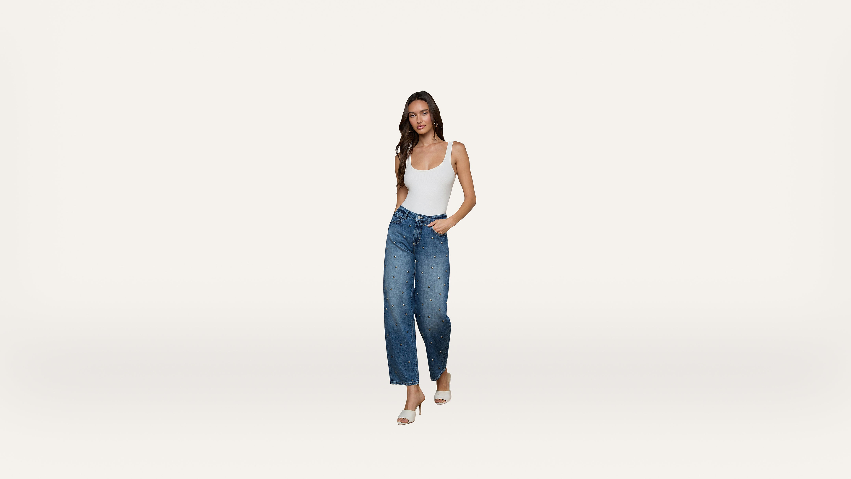 A woman wearing a white tank top, blue high-waisted wide-leg jeans with embellishments, and white heels stands against a plain white background.