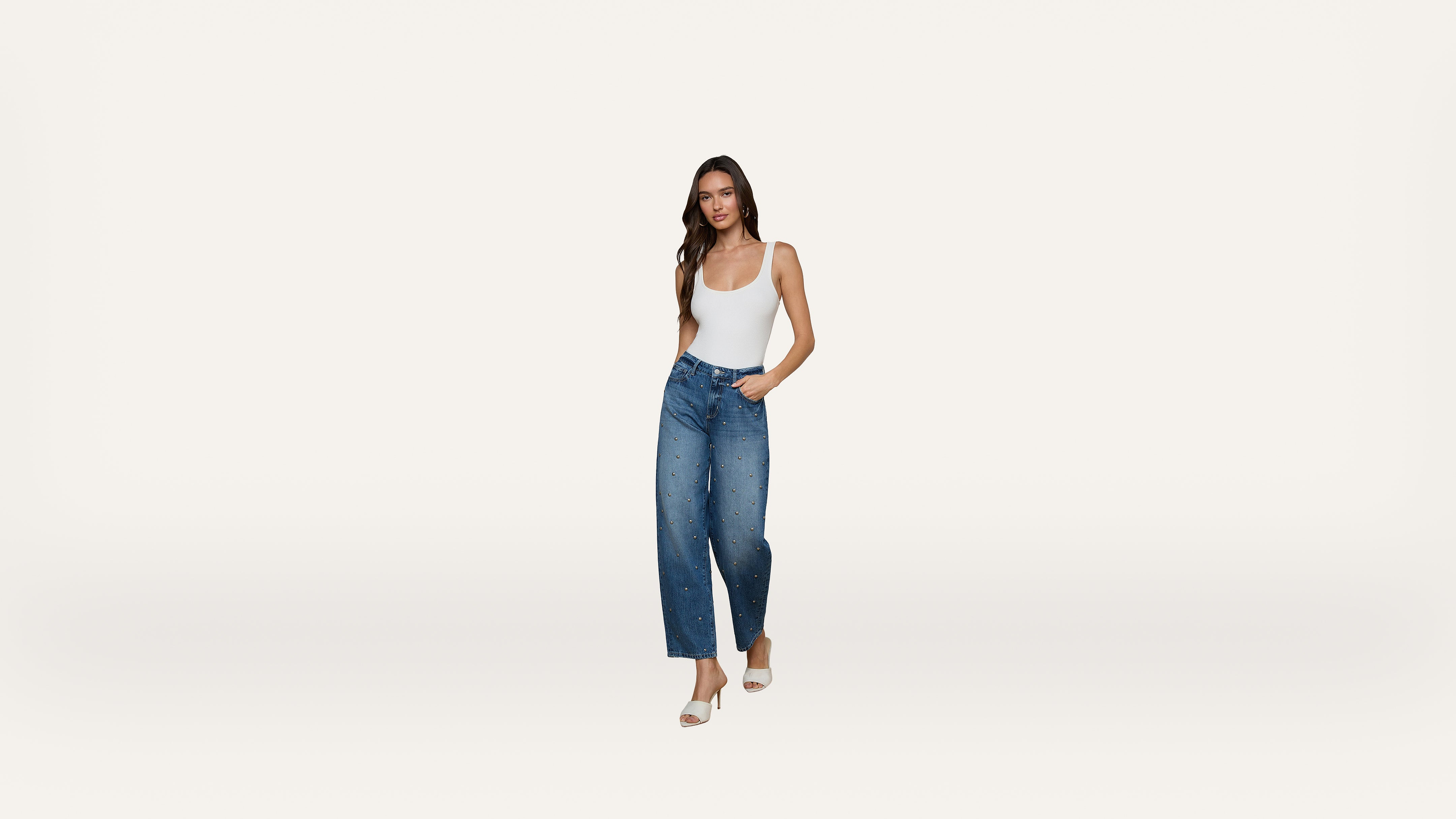 A woman wearing a white tank top, blue high-waisted wide-leg jeans with embellishments, and white heels stands against a plain white background.