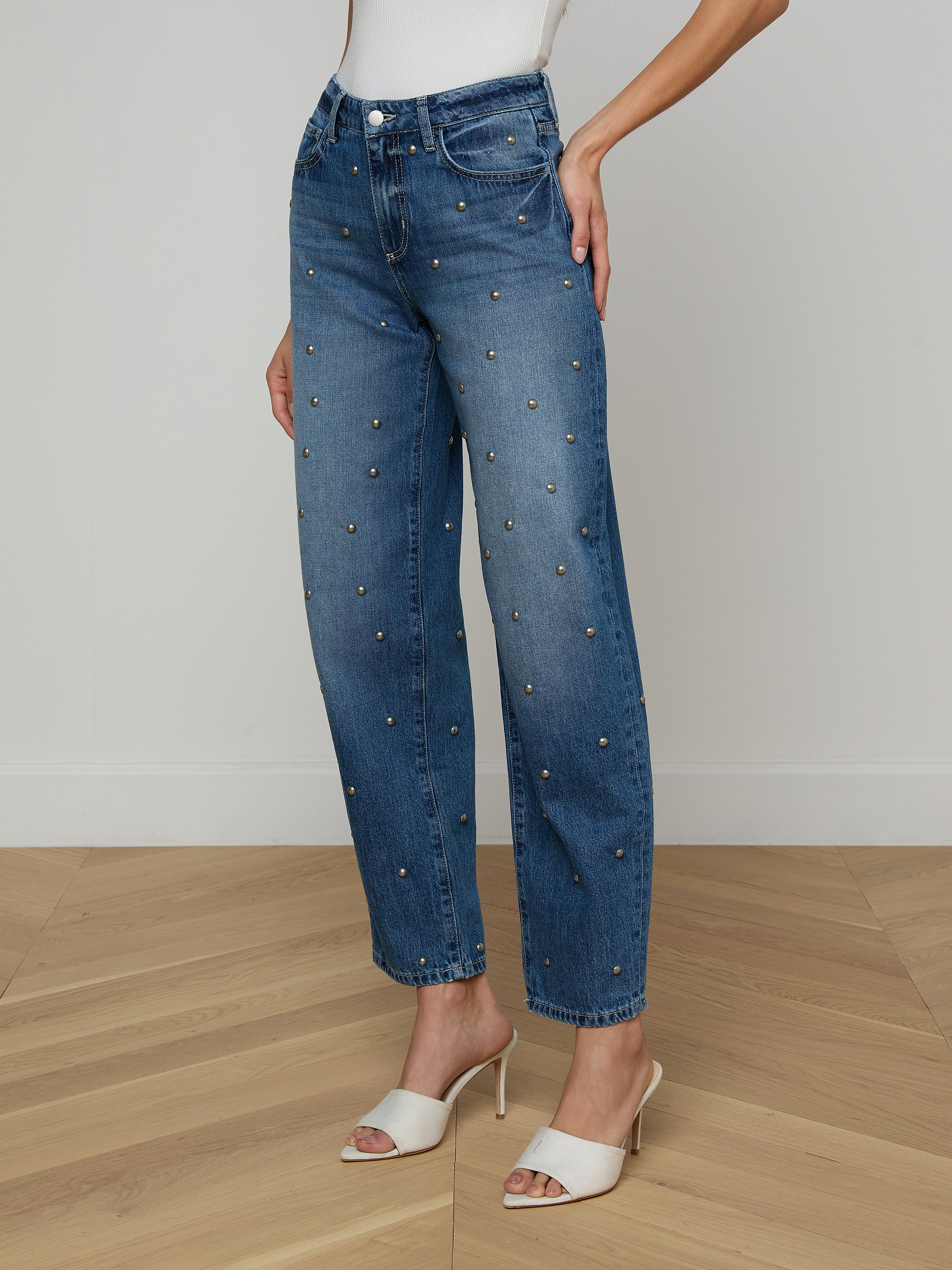 A person is wearing the Bex Studded Rodeo Jean, a pair of high rise blue jeans with small embellishments, styled with a white top and white open-toe heeled sandals, posed against a plain light background.