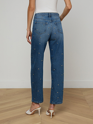 A person wears blue Bex Studded Rodeo Jeans with small studs and a white sleeveless top, standing on a light wood floor facing away from the camera. Their outfit is finished with white heeled sandals.