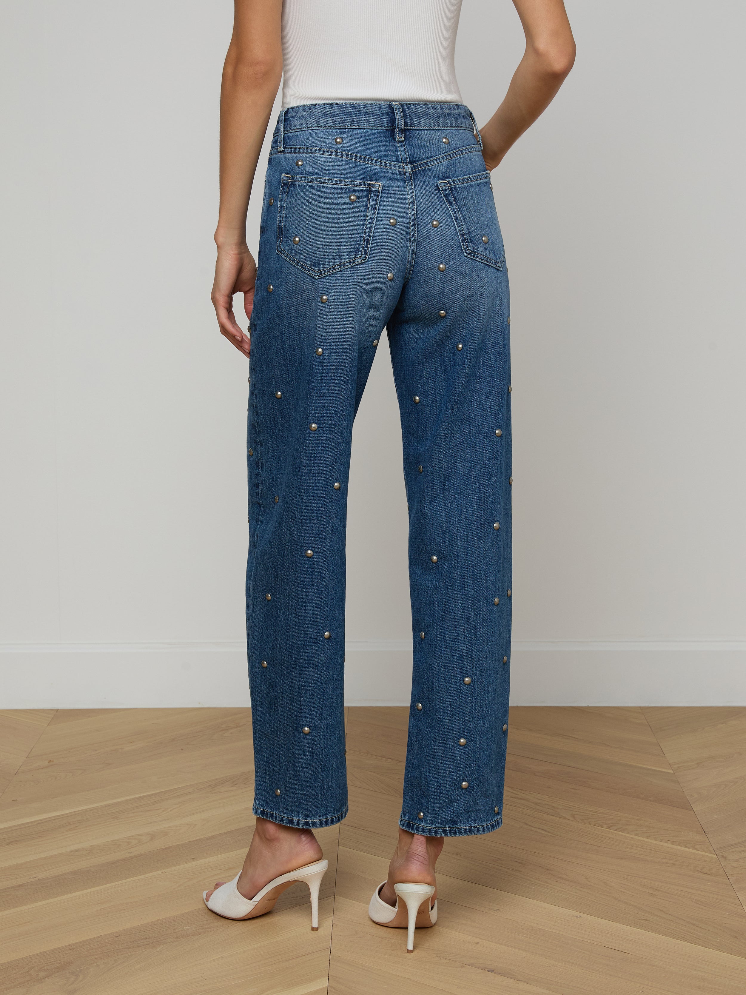 A person wearing a white sleeveless top, the Bex Studded Rodeo Jean in blue with small embellishments, and white high-heeled shoes is shown from the back against a plain background.