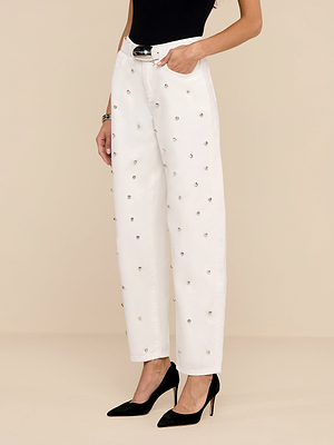 Una persona in piedi su uno sfondo beige indossa il Bex Embellished Rodeo Jean con cintura nera, top nero senza maniche e tacchi neri a punta, con una mano in tasca.