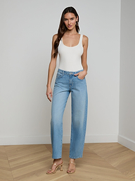 A woman stands against a plain white background wearing a white tank top, the Bex Rodeo Jean in light blue high-waisted barrel leg all-cotton denim, and beige heeled sandals.