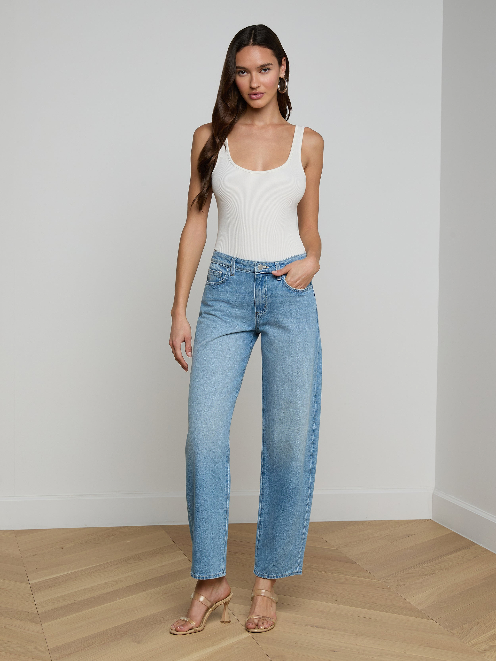 A woman stands against a plain white background wearing a white tank top, the Bex Rodeo Jean in light blue high-waisted barrel leg all-cotton denim, and beige heeled sandals.