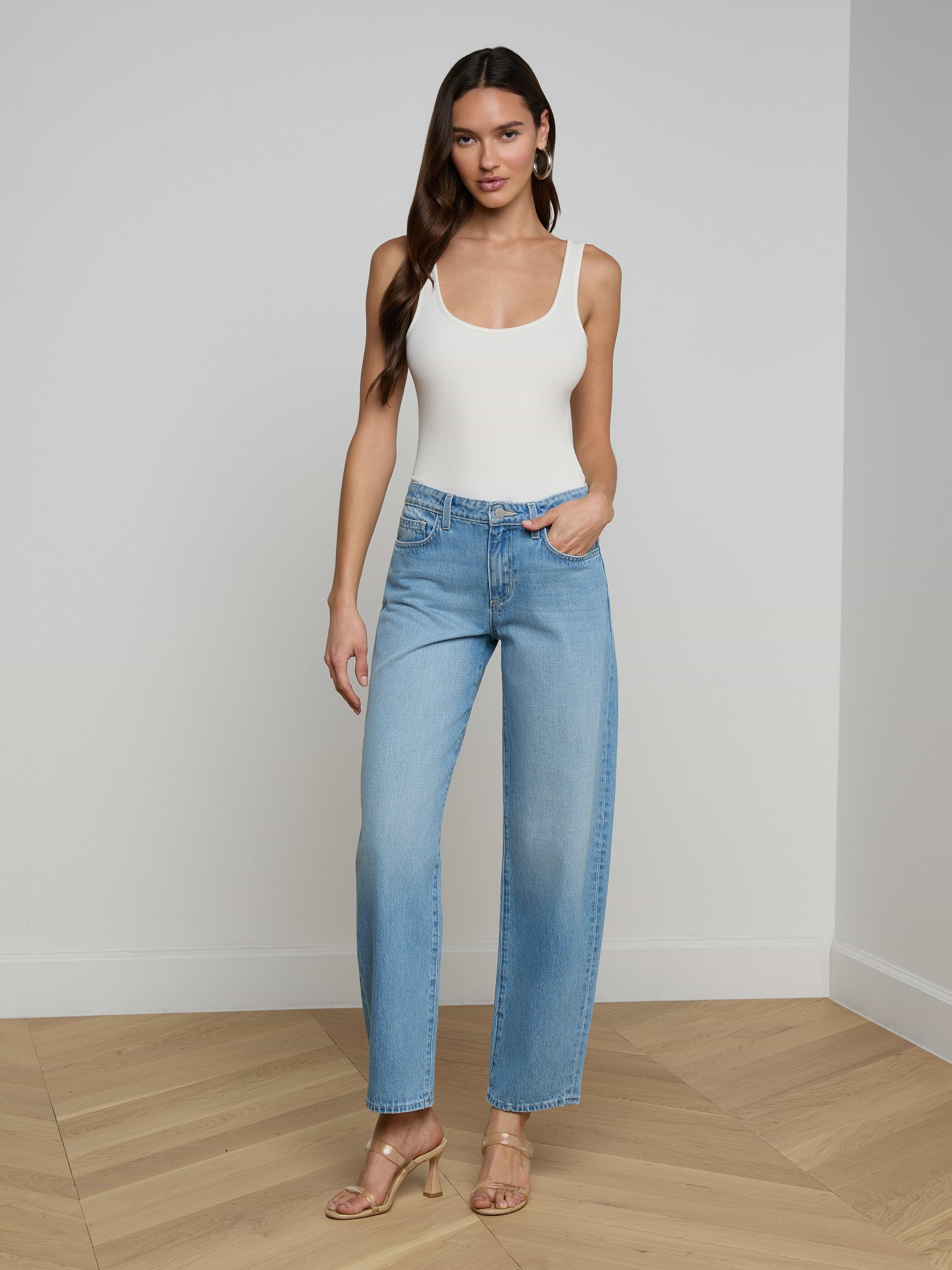 A woman stands against a plain white background wearing a white tank top, the Bex Rodeo Jean in light blue high-waisted barrel leg all-cotton denim, and beige heeled sandals.