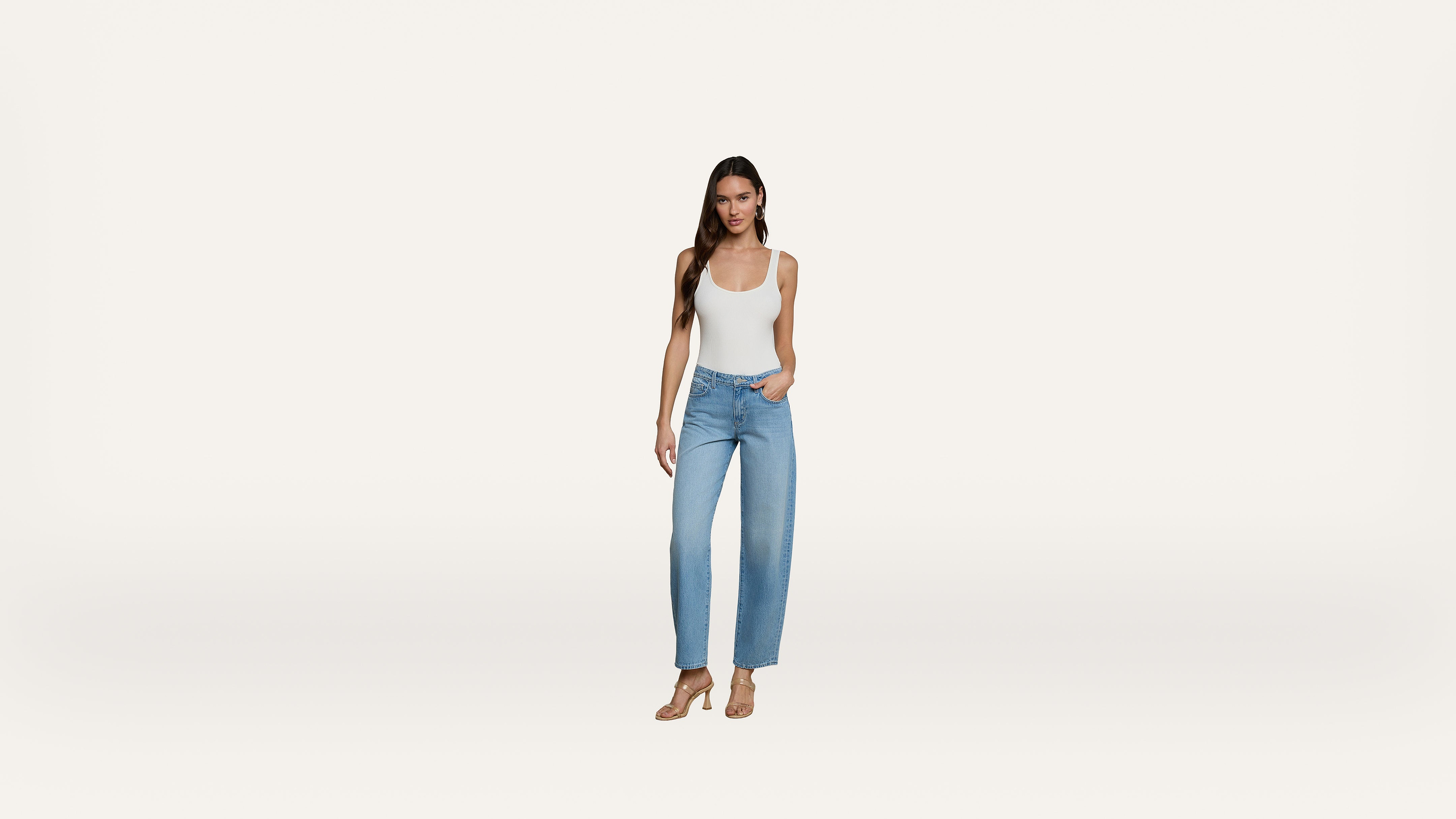 A woman stands against a white background wearing a white tank top, blue jeans, and nude heels, with one hand in her pocket.