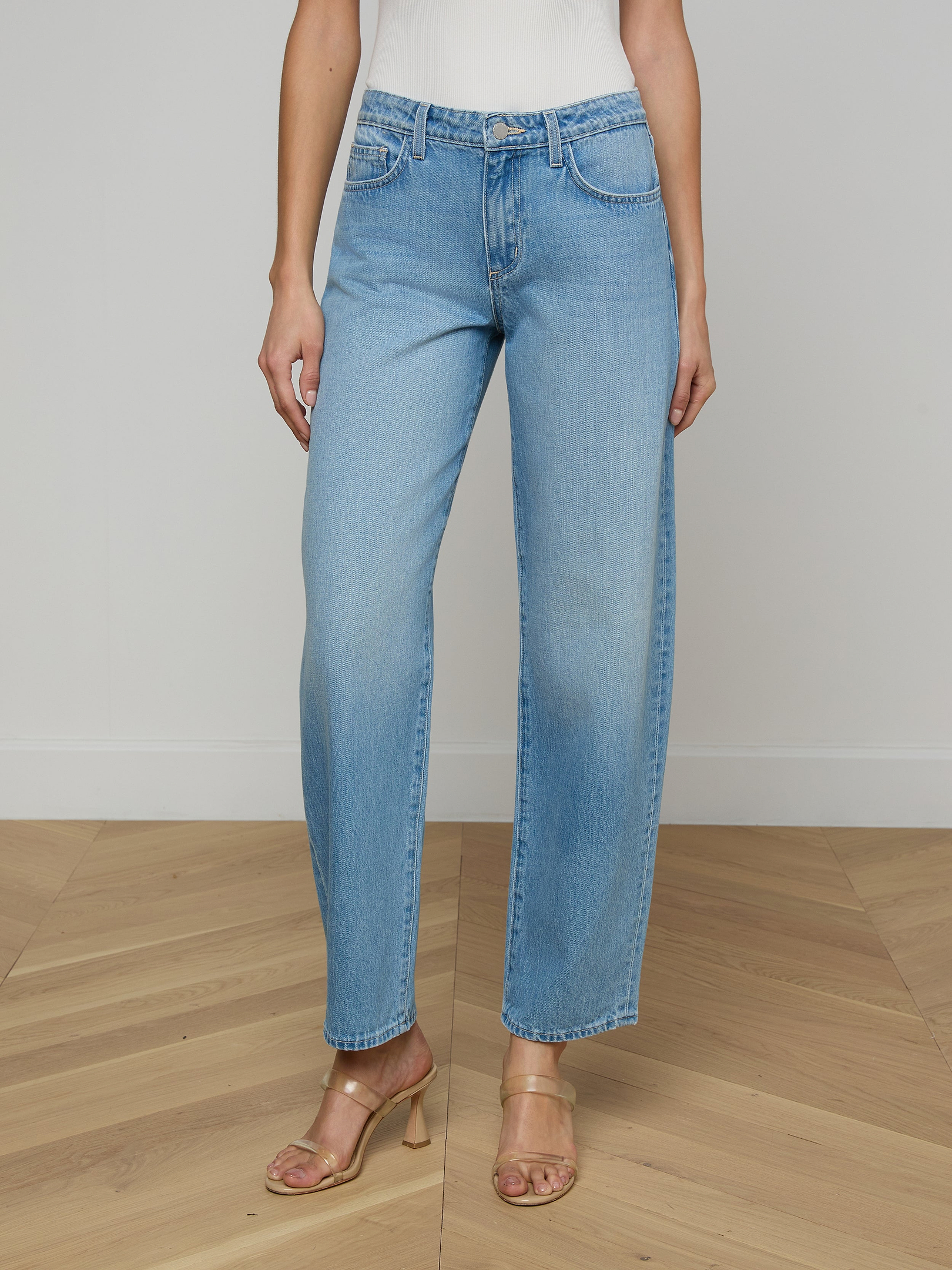 A person wearing the Bex Rodeo Jean in light blue all-cotton denim, paired with a white top and beige heeled sandals, standing against a plain light background.