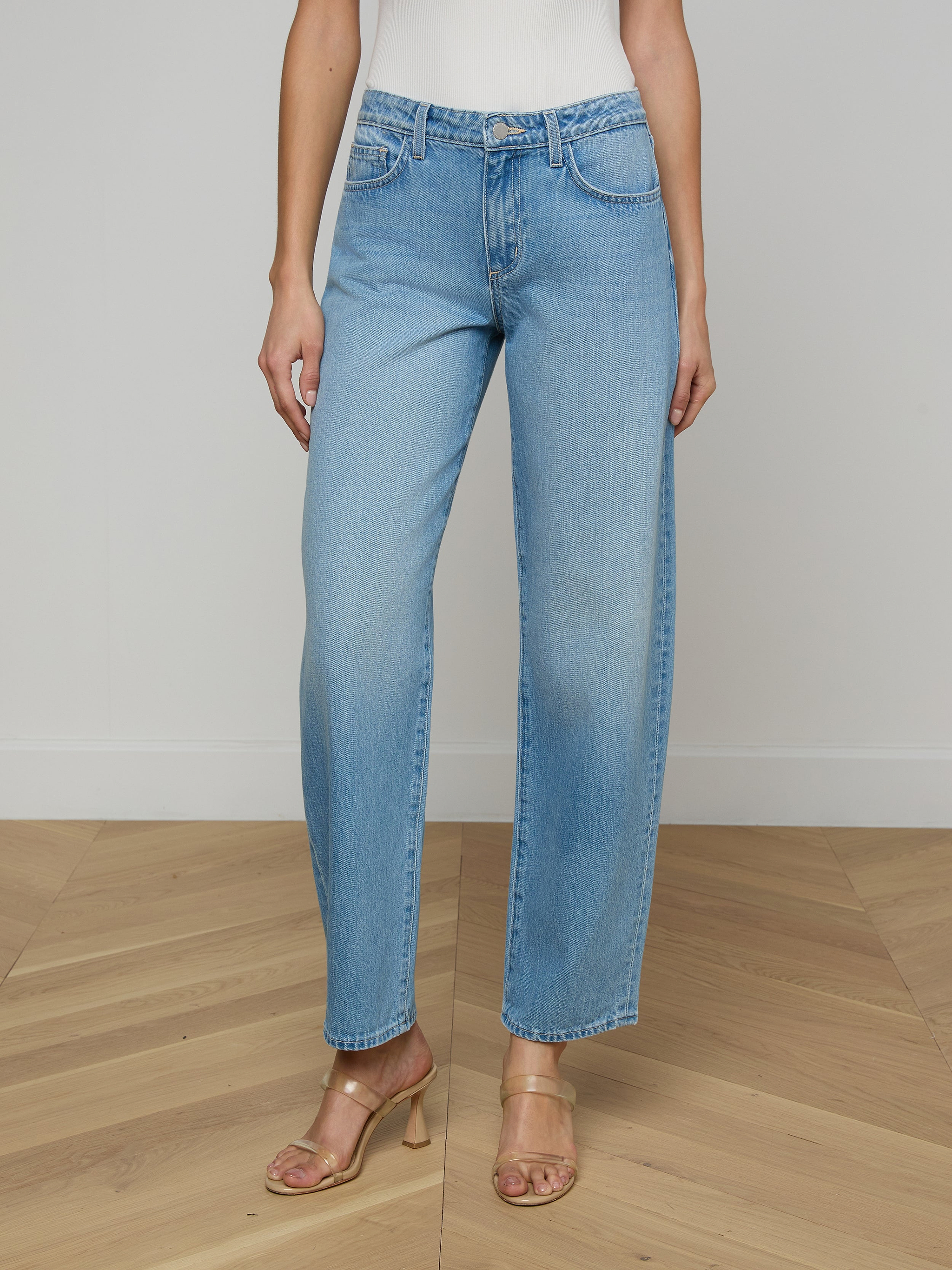 A person wearing the Bex Rodeo Jean in light blue all-cotton denim, paired with a white top and beige heeled sandals, standing against a plain light background.