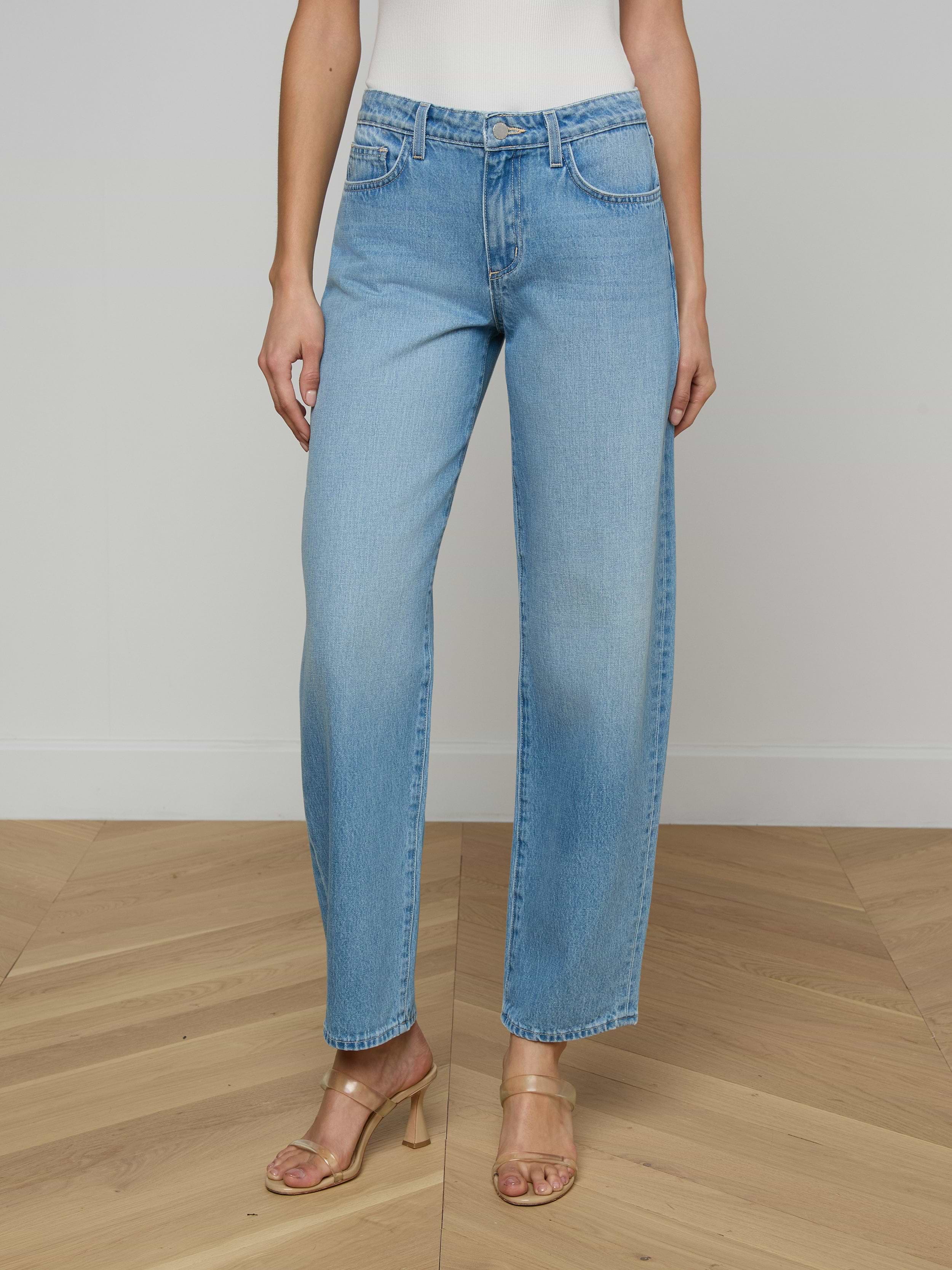 A person wearing the Bex Rodeo Jean in light blue all-cotton denim, paired with a white top and beige heeled sandals, standing against a plain light background.