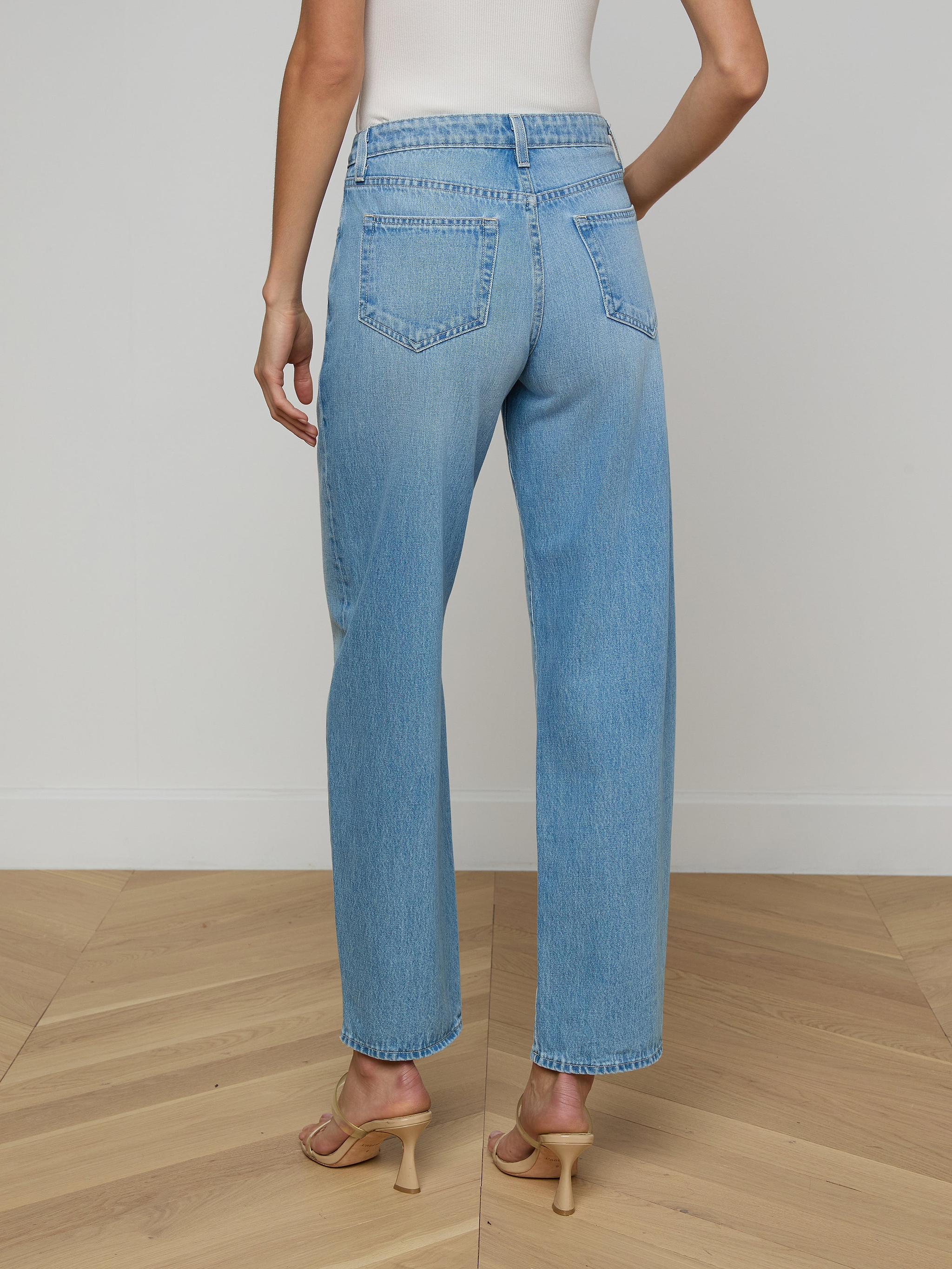 A person is shown from the back against a plain, light background wearing the Bex Rodeo Jean—a pair of light blue, high-waisted barrel leg jeans crafted from all-cotton denim—paired with heeled sandals.