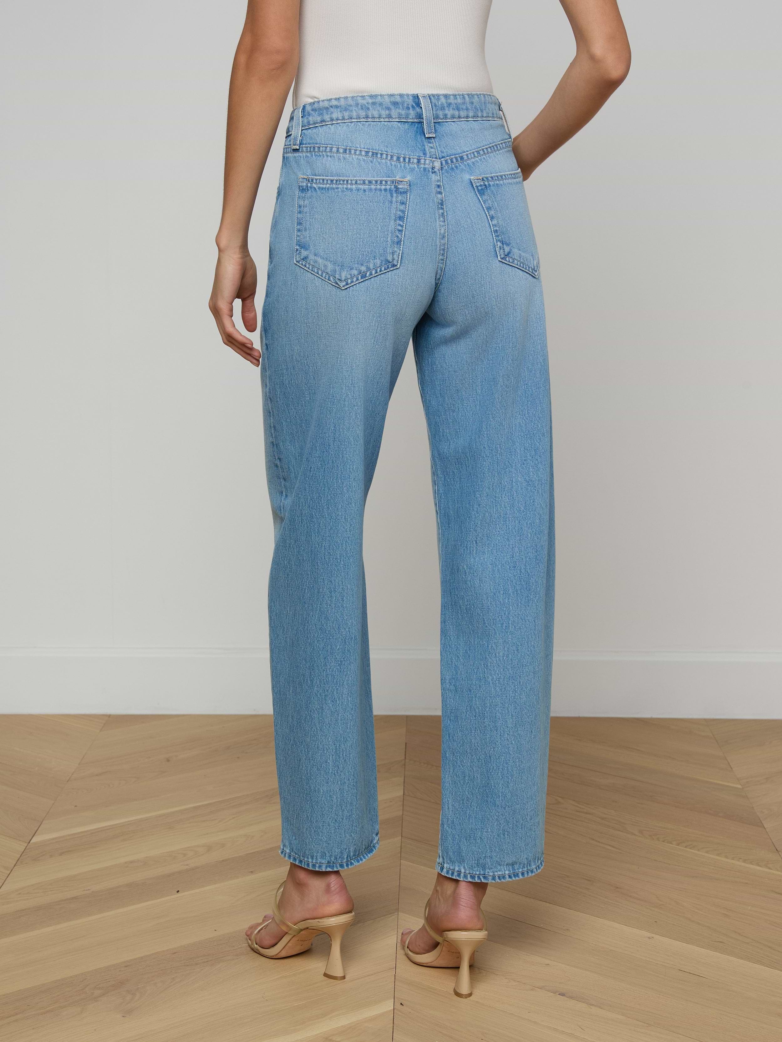 A person is shown from the back against a plain, light background wearing the Bex Rodeo Jean—a pair of light blue, high-waisted barrel leg jeans crafted from all-cotton denim—paired with heeled sandals.