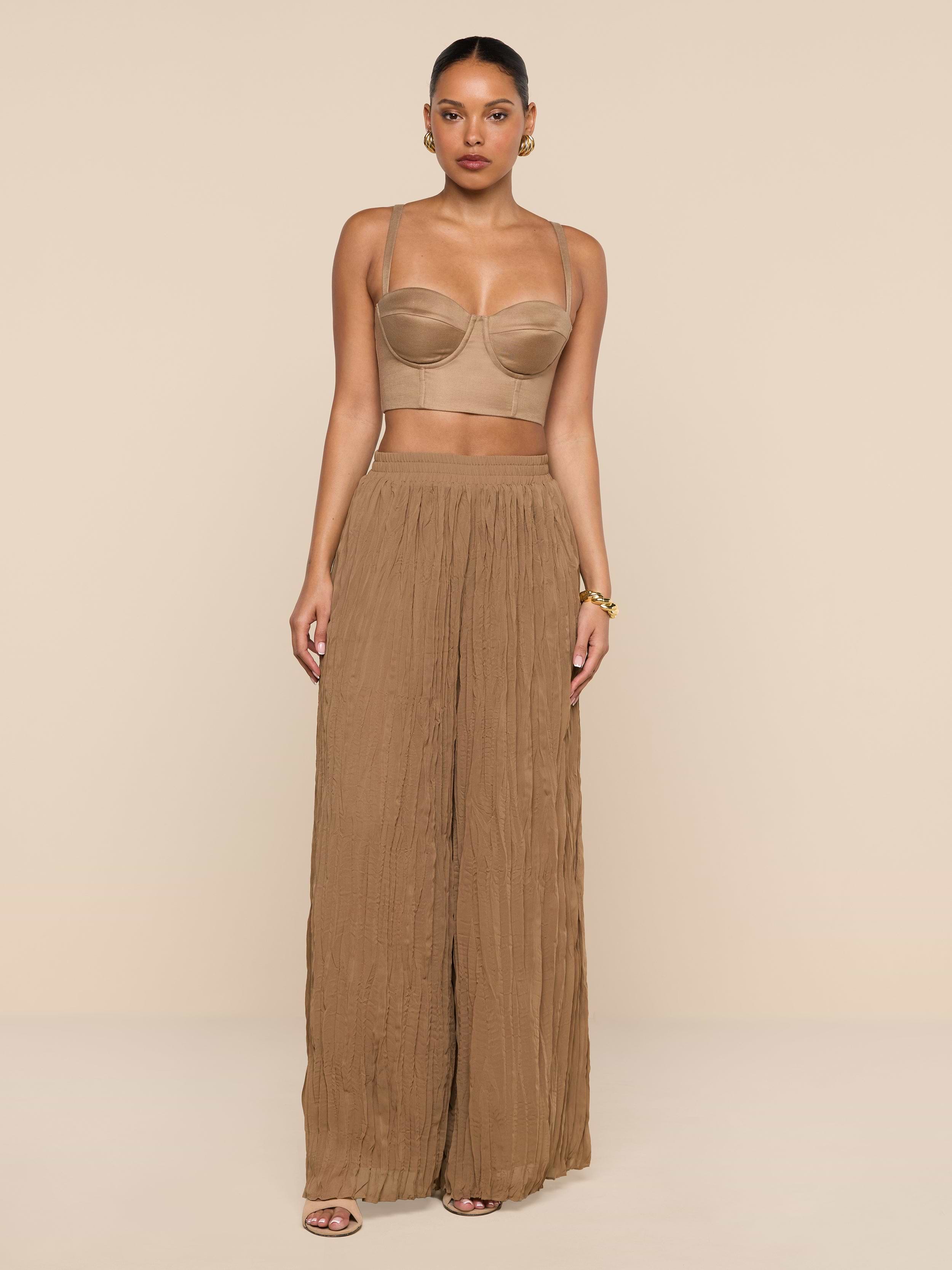 A woman stands against a plain white background wearing a tan bandeau top, the Ethel Pleated Pant—a sheer, wide-legged brown summer style—sandals, and gold hoop earrings. Her hair is pulled back and her hands rest relaxed at her sides.
