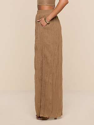 Wearing the Ethel Pleated Pant in light brown crinkle chiffon with a matching top, a woman stands poised against a beige background, her hand in her pocket and a chunky gold bracelet on her wrist.