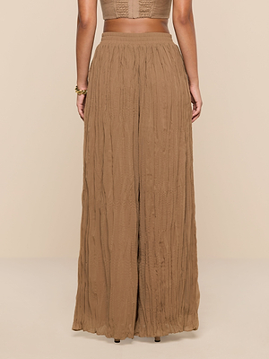 A person stands facing away, wearing the Ethel Pleated Pant—high-waisted, wide-leg, light brown crinkle chiffon made of flowy fabric. The background is solid beige.