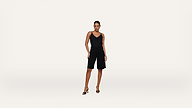 A woman stands against a plain white background, wearing a black sleeveless top, black knee-length shorts, and black high-heeled sandals. She poses confidently with one hand on her hip.