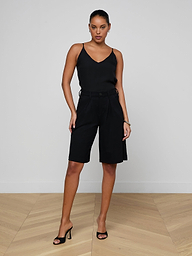 A woman stands confidently against a plain white background, wearing a black sleeveless top, the Akasha Bermuda Short in high-rise black stretch ponte, and black heeled sandals.