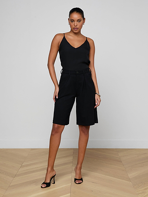 A woman poses confidently in a minimalist room, facing the camera, wearing the Akasha Bermuda Short with a black stretch ponte sleeveless top and black high-heeled sandals. Her hair is pulled back.