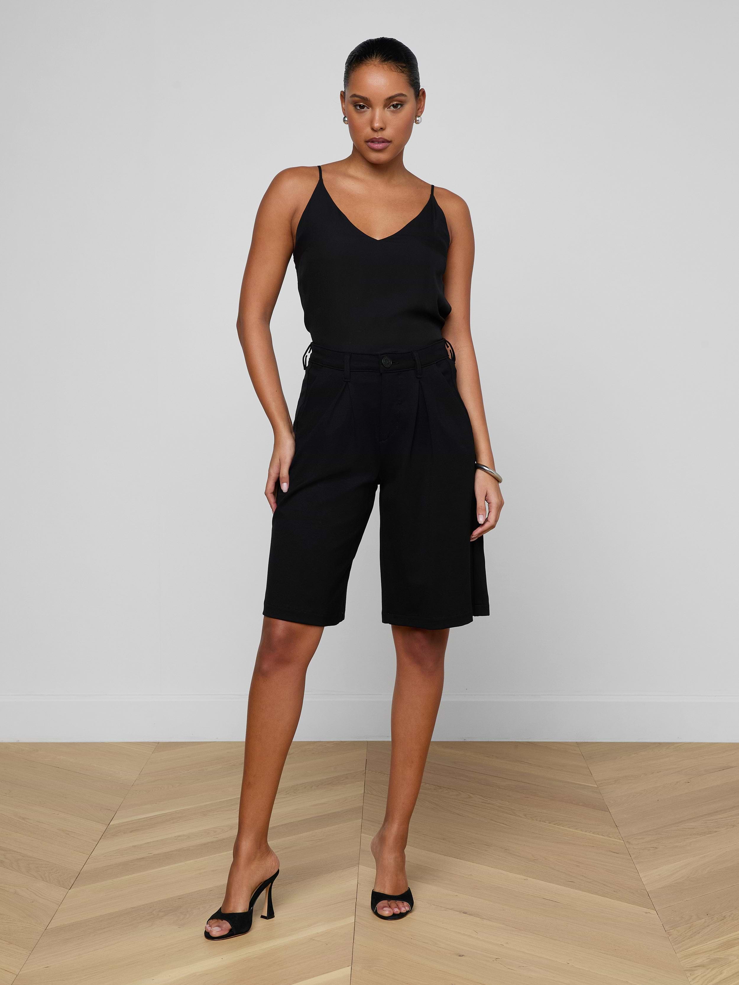 A woman stands confidently against a plain white background, wearing a black sleeveless top, the Akasha Bermuda Short in high-rise black stretch ponte, and black heeled sandals.