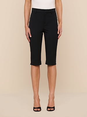 Someone wears the Leni Pedal Pusher black knee-length tailored shorts in stretch suit fabric, paired with a white sleeveless top and black open-toe heels, standing against a neutral beige background.