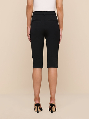 A person is seen from behind against a beige background, wearing the Leni Pedal Pusher in black stretch suit fabric and black heeled sandals.