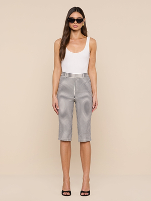 A woman stands against a beige background wearing a white sleeveless top, the Leni Pedal Pusher in black and white striped cotton seersucker shorts, black high-heeled sandals, and large black sunglasses.