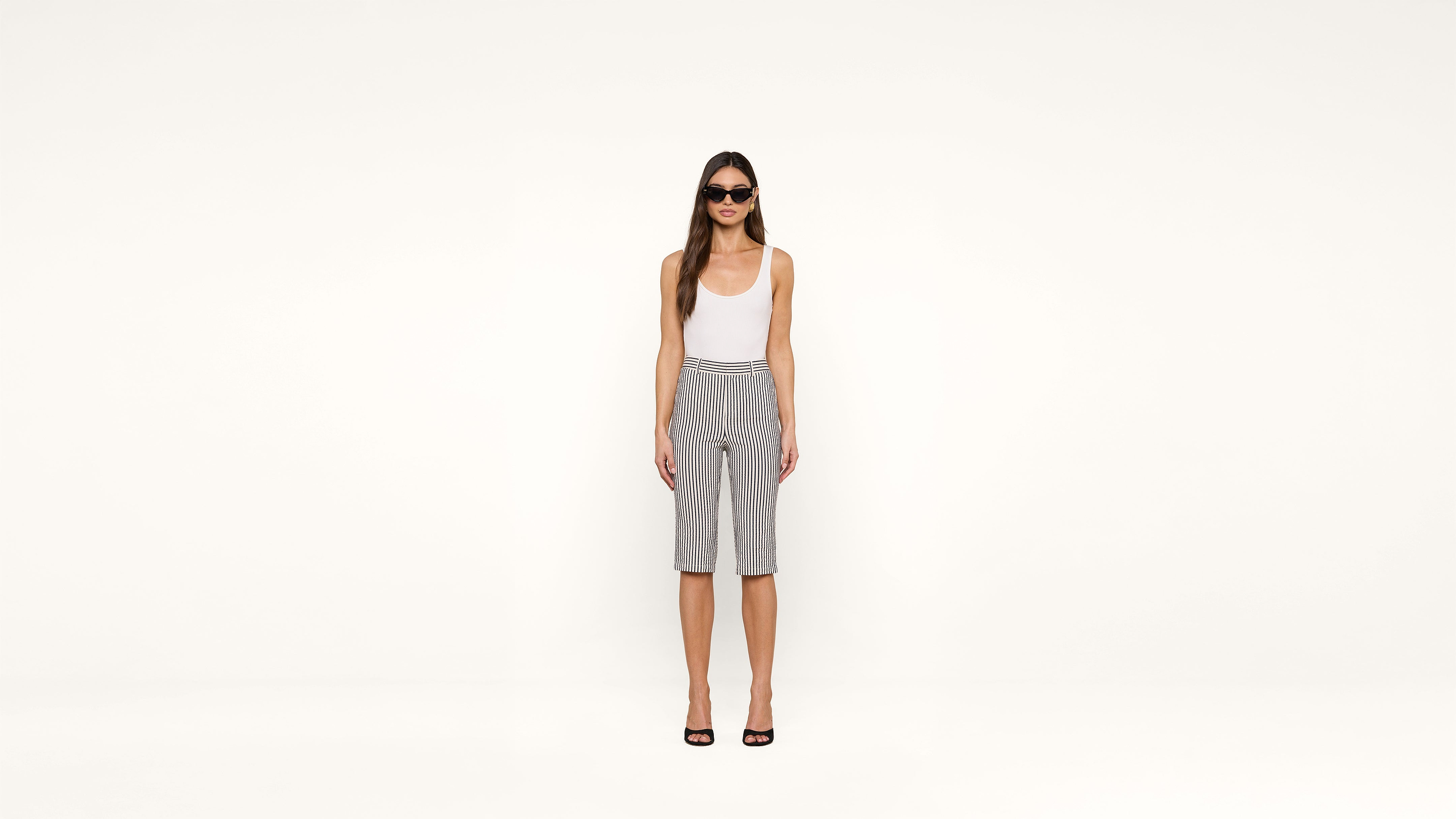 Woman stands against a plain white background, wearing sunglasses, a white tank top, black and white striped knee-length shorts, and black open-toe heels.