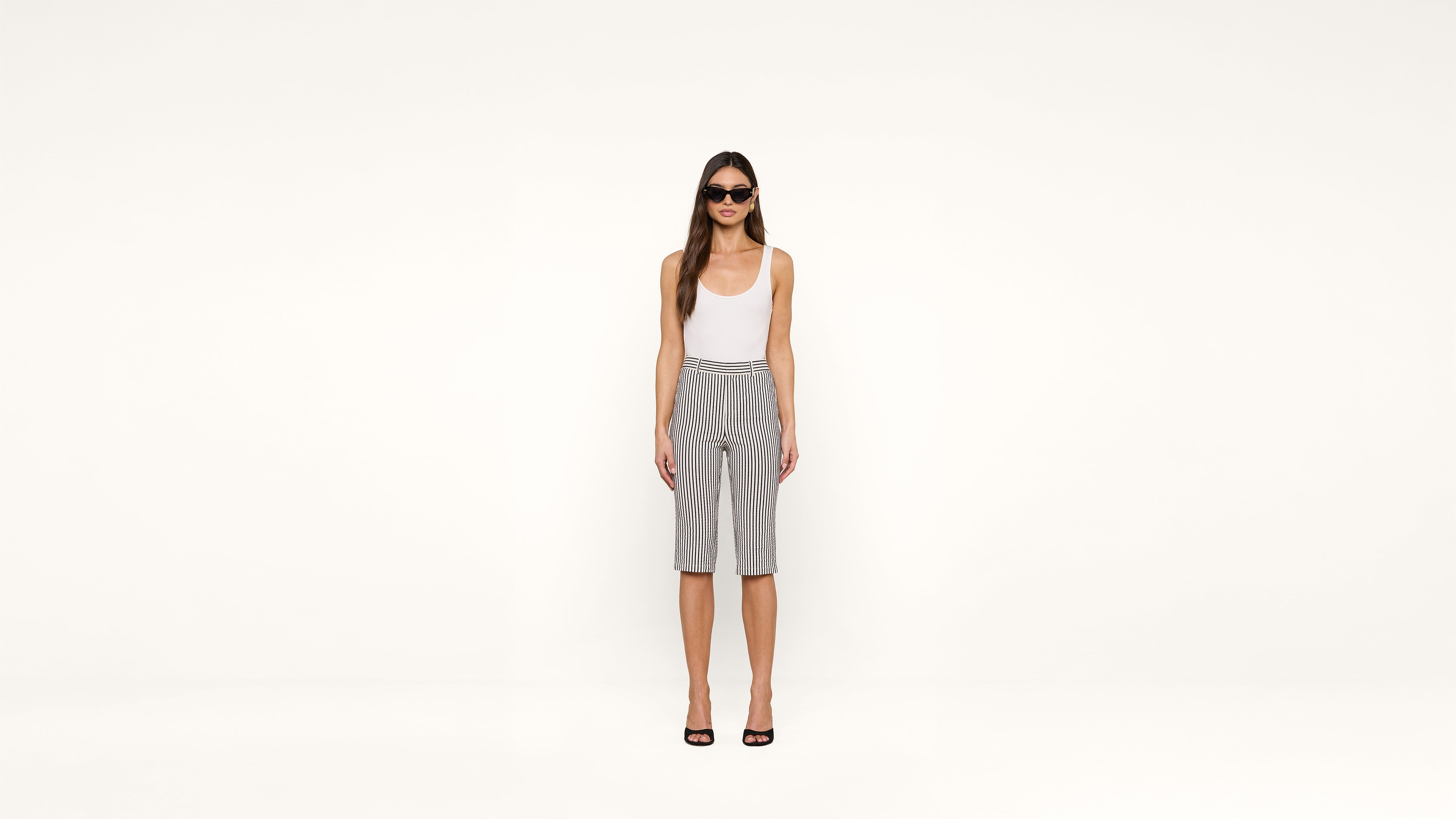 Woman stands against a plain white background, wearing sunglasses, a white tank top, black and white striped knee-length shorts, and black open-toe heels.