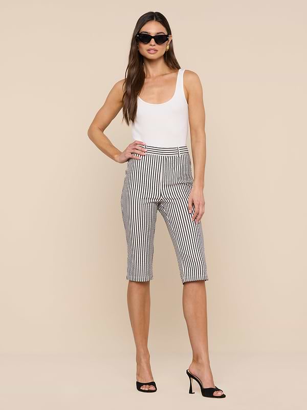 A woman stands confidently against a plain white background, wearing a white tank top, the Leni Pedal Pusher in black-and-white striped cotton seersucker, black high heels, and sunglasses.