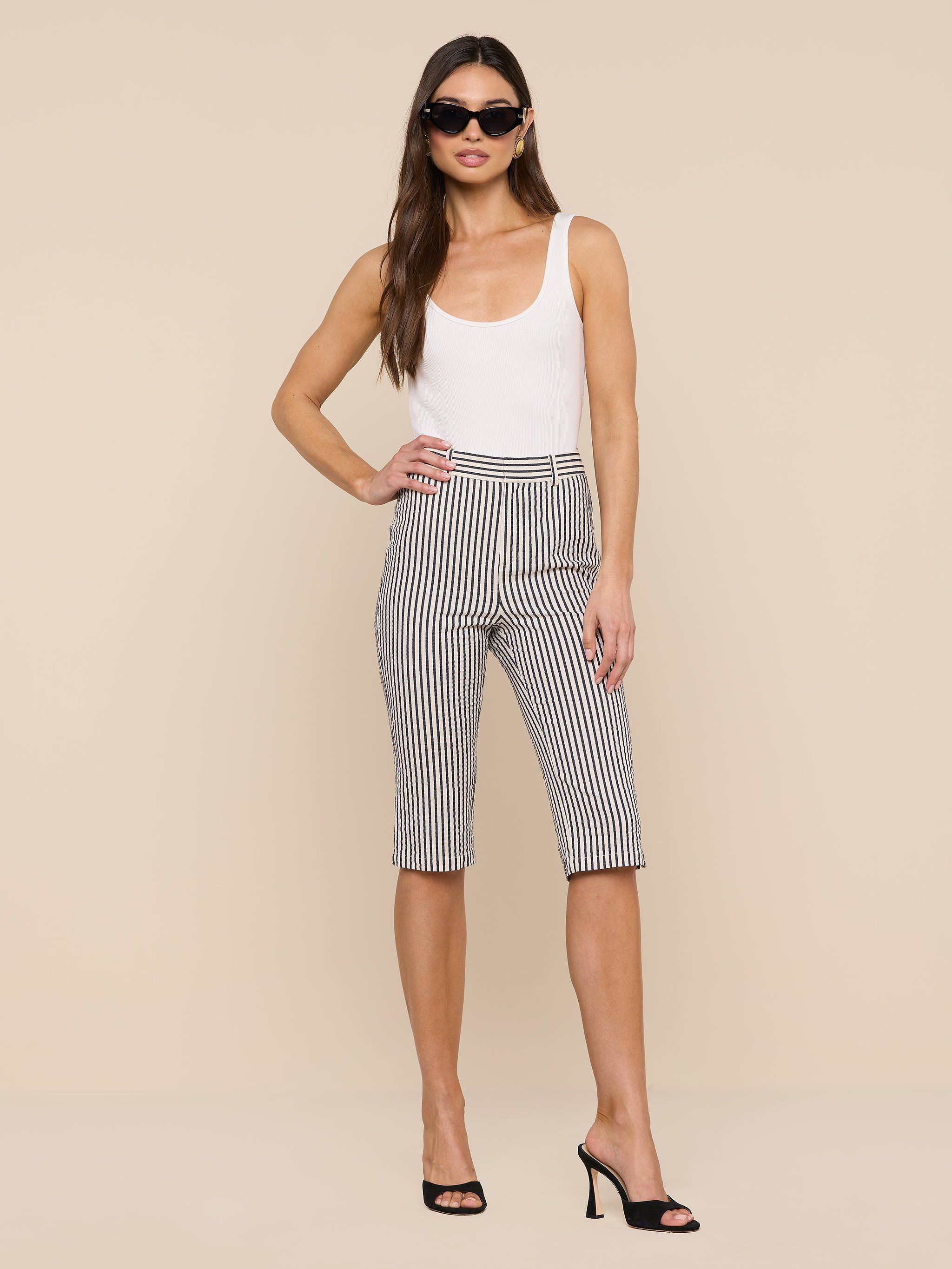 A woman stands confidently against a plain white background, wearing a white tank top, the Leni Pedal Pusher in black-and-white striped cotton seersucker, black high heels, and sunglasses.