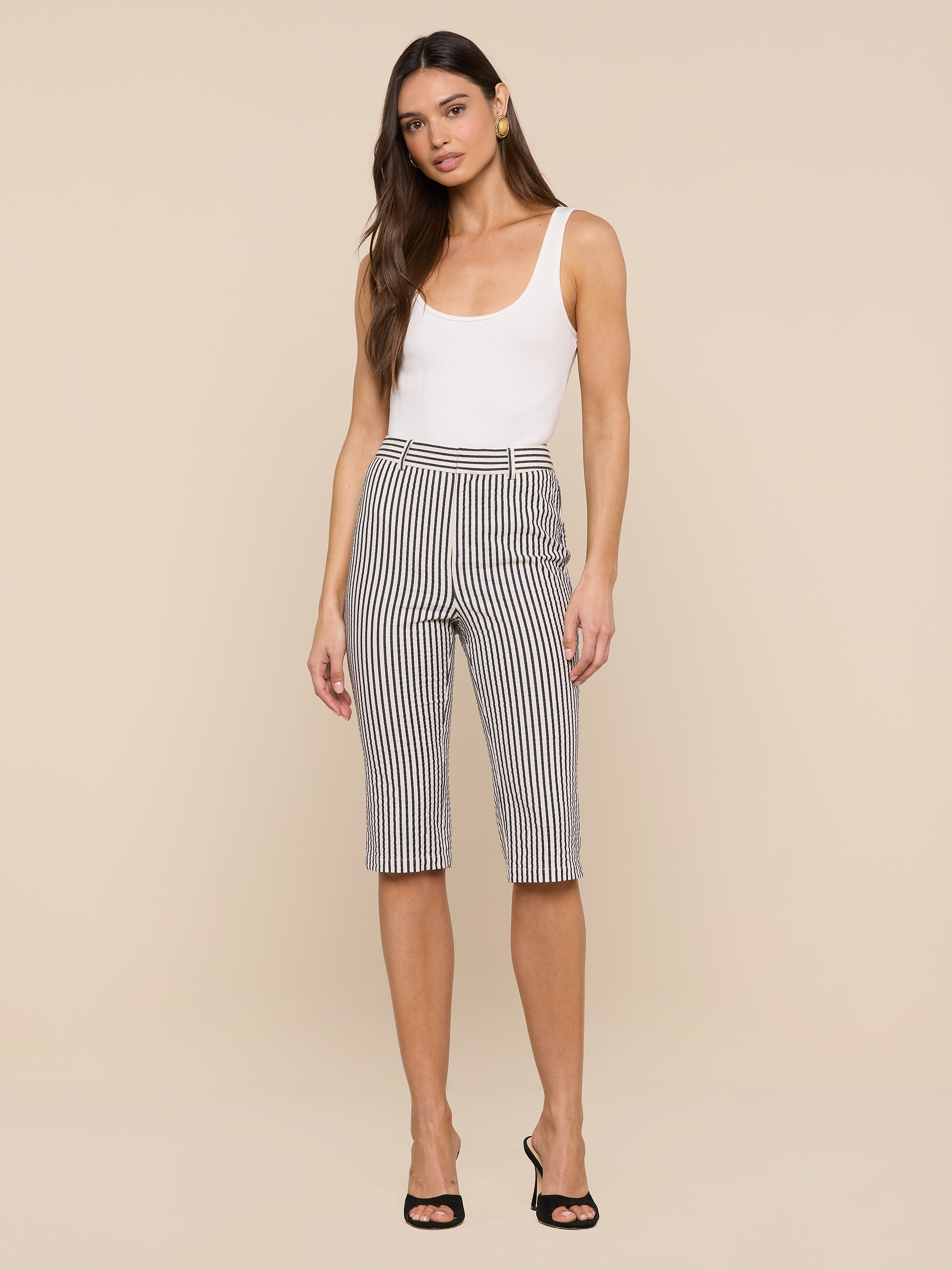 A woman stands against a white background, wearing a white tank top, the Leni Pedal Pusher in black and white vertical striped cotton seersucker, and black open-toe heels.