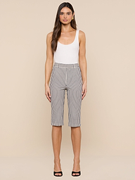 A woman stands against a plain white background, wearing a white tank top, the Leni Pedal Pusher high-waisted knee-length black-and-white cotton seersucker shorts, and black open-toe heels.