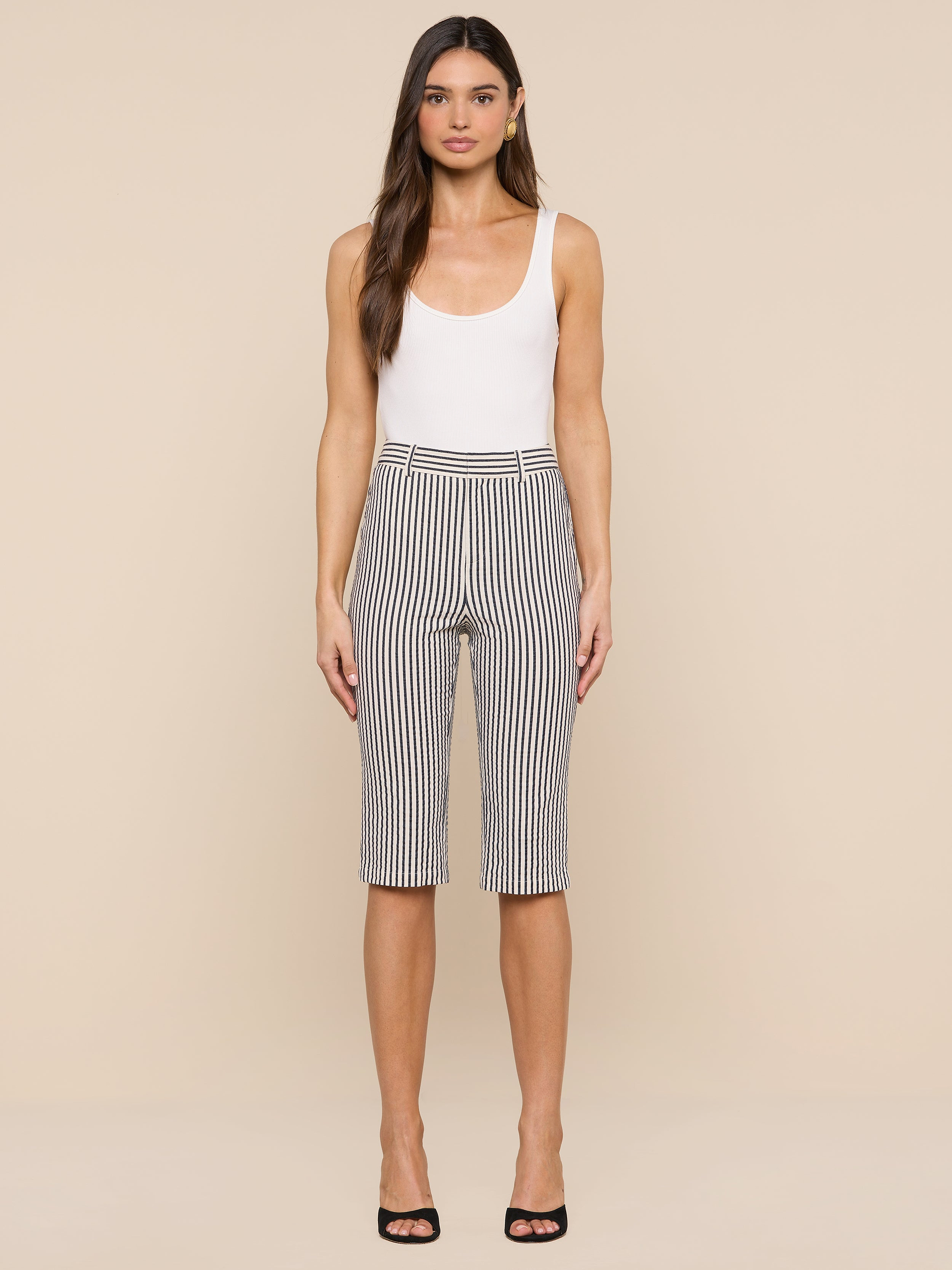 A woman stands against a plain white background, wearing a white tank top, the Leni Pedal Pusher high-waisted knee-length black-and-white cotton seersucker shorts, and black open-toe heels.