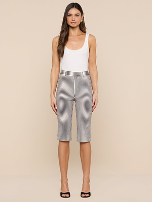 A woman stands against a beige background, wearing a white sleeveless top, black high-heeled shoes, and the Leni Pedal Pusher: black and white vertically striped cotton seersucker shorts.