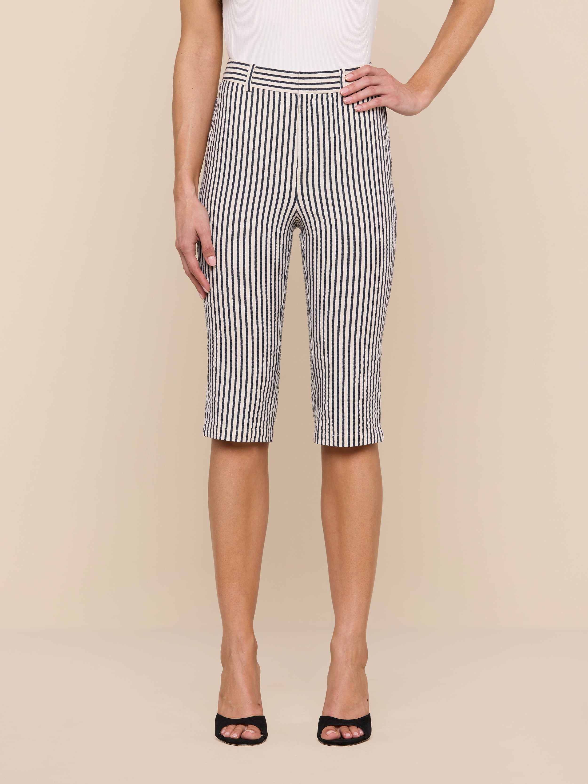 A person stands against a plain background, showcasing the lower half of their body dressed in the Leni Pedal Pusher—black and white vertically striped cotton seersucker knee-length shorts—paired with a sleeveless white top and black open-toe heels.