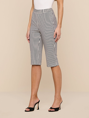 A woman wearing the Leni Pedal Pusher black and white striped cotton seersucker Bermuda shorts with a white sleeveless top and black high-heeled sandals, standing against a beige background.