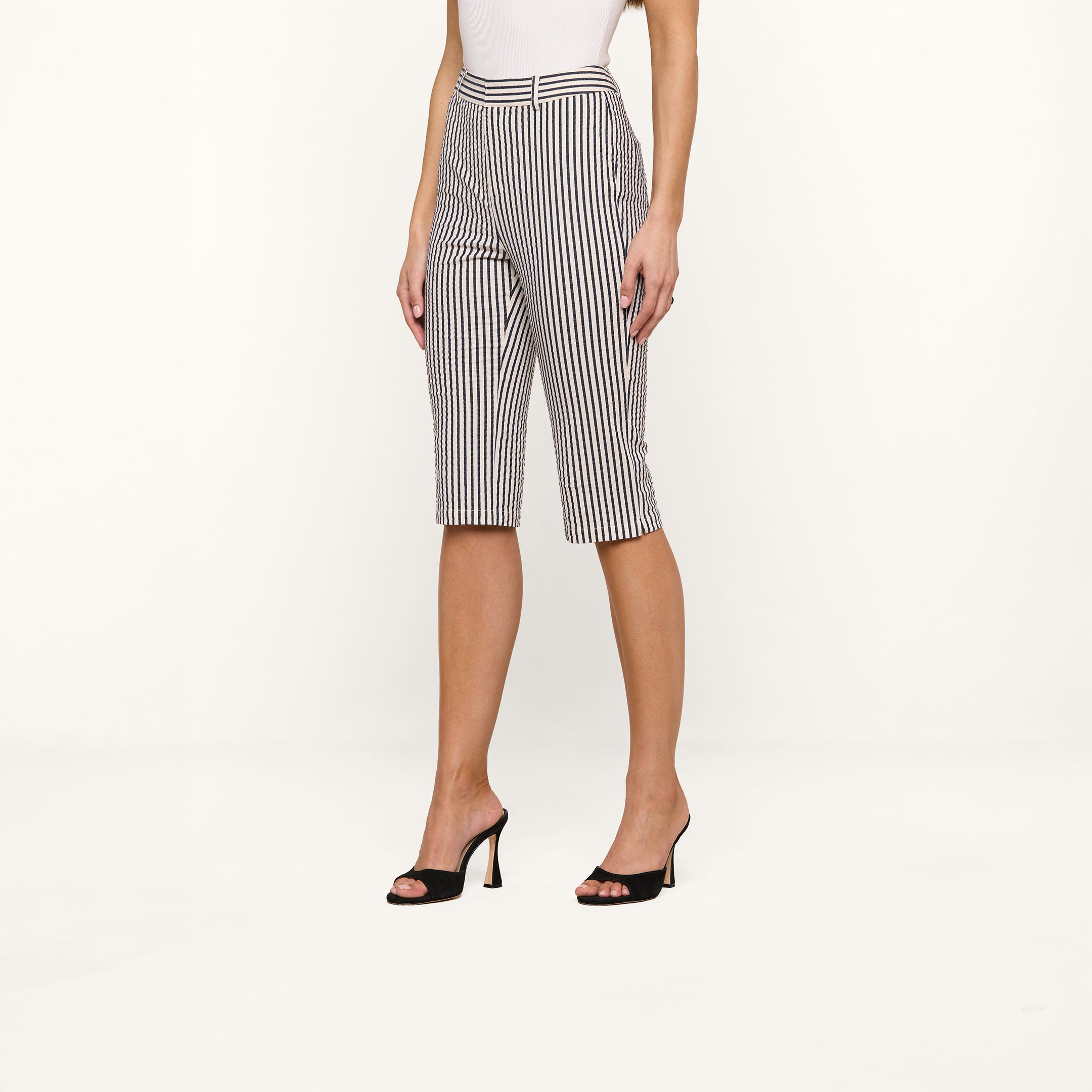 A woman stands against a white background wearing the Leni Pedal Pusher—black and white vertical striped cotton seersucker knee-length shorts—paired with a white top and black high-heeled sandals. Her arms rest by her sides, and her face is not visible.