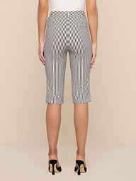 A person stands facing away against a plain white background, wearing the Leni Pedal Pusher—knee-length black and white vertical striped cotton seersucker shorts—paired with a white top and black open-heeled shoes.