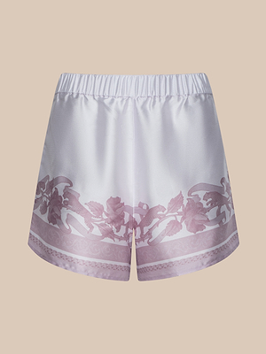 The Maela Short is a silky white pull-on style with a light purple floral vine pattern along the bottom hem, crafted from shiny poly twill. It features an elastic waistband and is shown against a beige background.