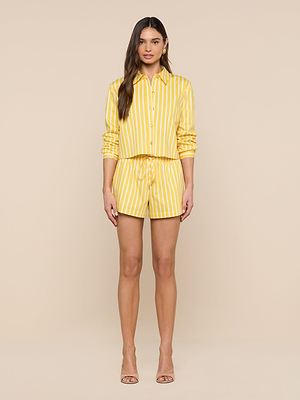 A woman stands against a beige background wearing the Royce Poplin Short and a matching yellow-and-white striped button-up shirt with nude heels, facing forward with her arms relaxed at her sides.