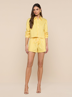 A woman stands against a beige background wearing the Royce Poplin Short and a matching yellow and white vertically striped long-sleeve shirt, paired with nude heeled sandals.
