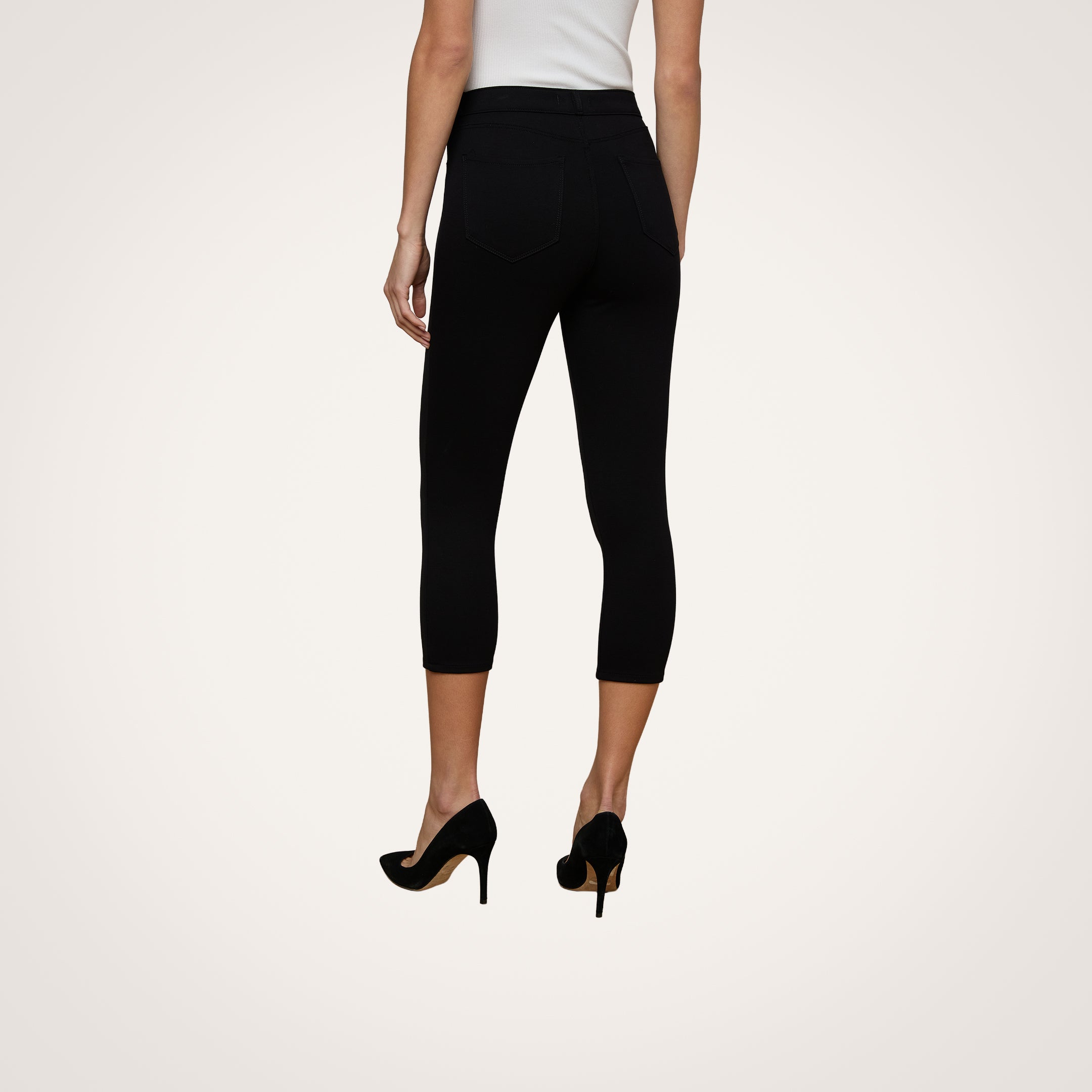 A person is seen from the back, standing against a plain light background, wearing a white sleeveless top and the Amaris Pedal Pusher—black ponte knit pants with a body-hugging fit—paired with black high heels.