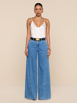 A woman stands against a beige background wearing the Niles Wide-Leg Jean in blue, paired with a white V-neck sleeveless top, a black belt with a gold buckle, gold hoop earrings, and a gold bracelet.