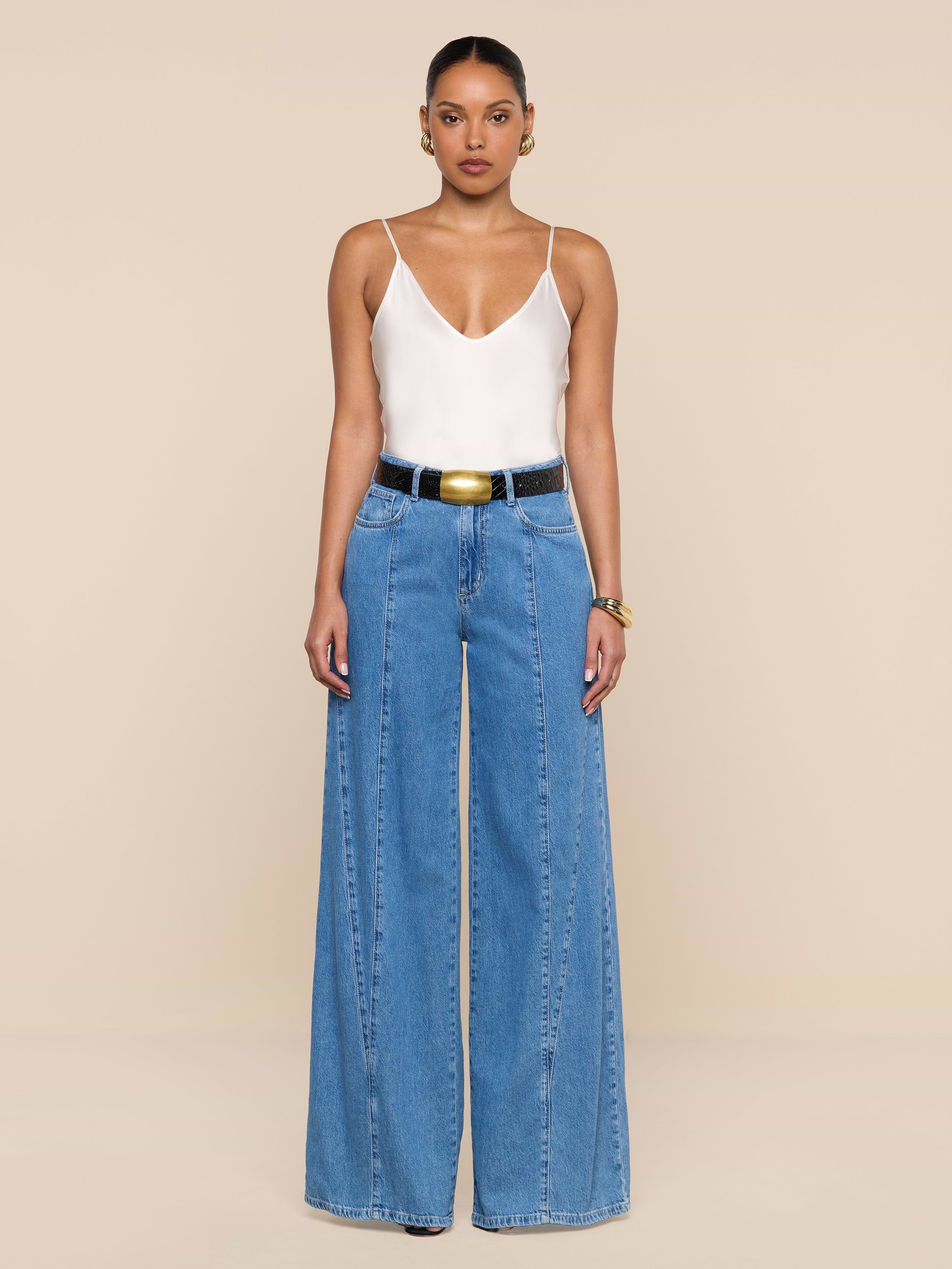 A person stands against a white background wearing a white camisole top, the Niles Wide-Leg Jean in a high rise lightweight denim, a black belt with a large gold buckle, and hoop earrings.