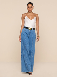 A woman stands against a plain white background wearing a white tank top, the Niles Wide-Leg Jean styled with a black belt, black open-toe heels, and gold jewelry.