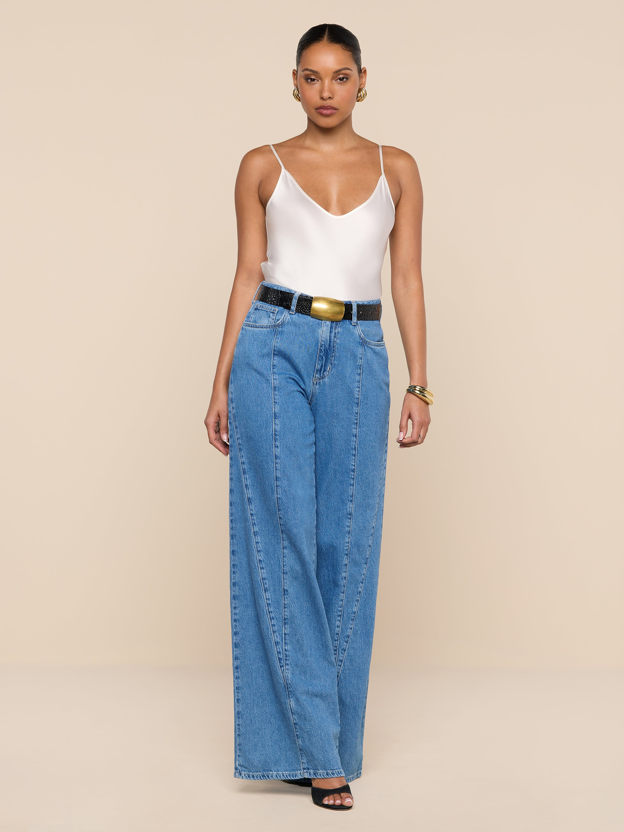 A woman stands against a plain white background wearing a white tank top, the Niles Wide-Leg Jean styled with a black belt, black open-toe heels, and gold jewelry.