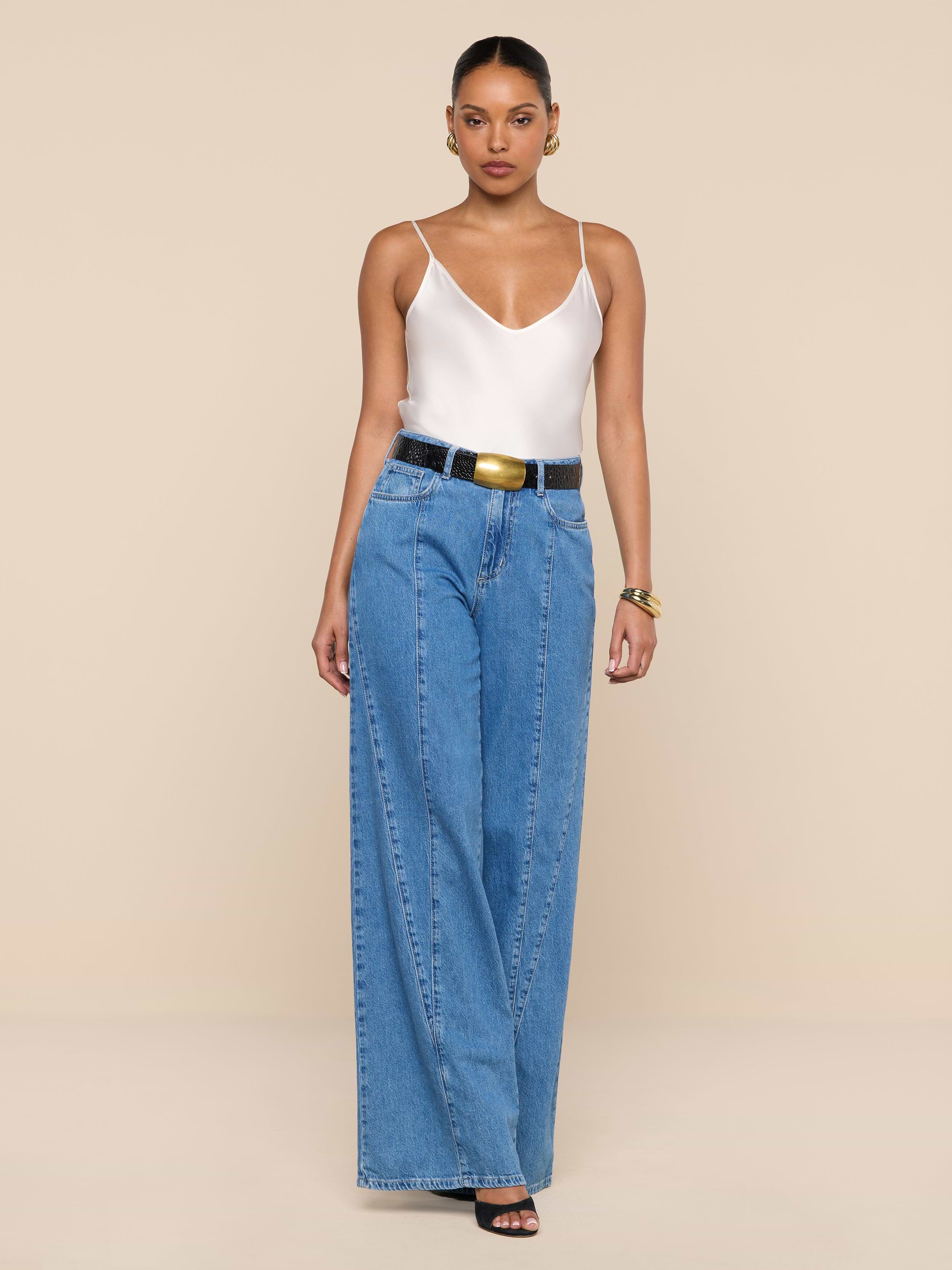 A woman stands against a plain white background wearing a white tank top, the Niles Wide-Leg Jean styled with a black belt, black open-toe heels, and gold jewelry.