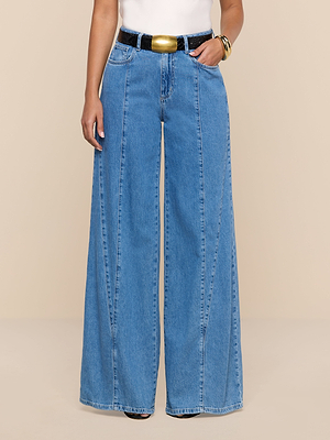 A person wearing Niles Wide-Leg Jeans in blue with vertical seams, paired with a black belt with a large gold buckle, a white top, and a gold bracelet, stands against a beige background.